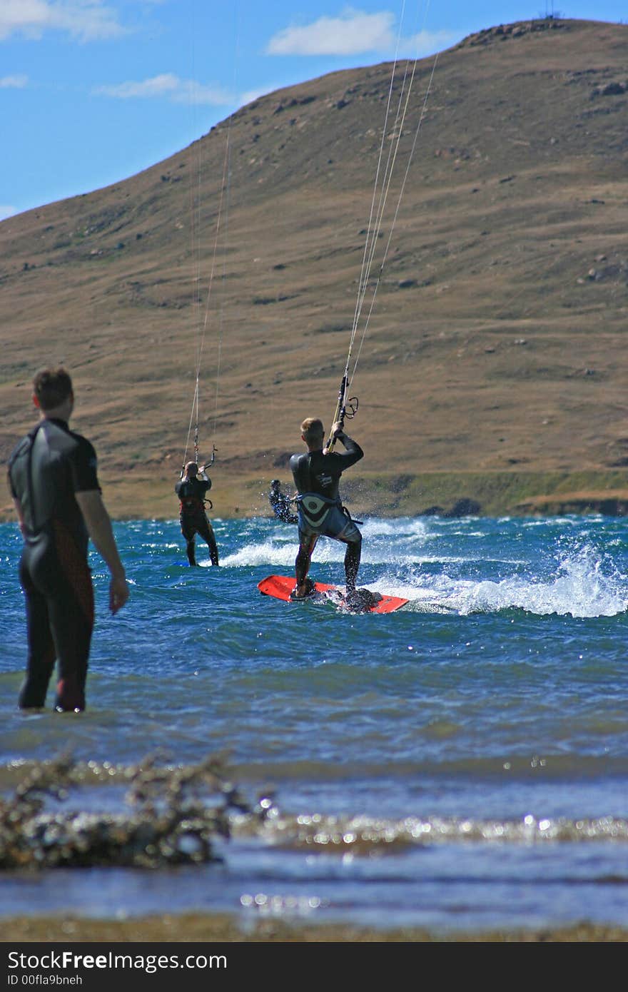 A kite surfer watching 3 other kitesurfers out in the water kitesurfing. All in wetsuits with scenic mountain behind. A kite surfer watching 3 other kitesurfers out in the water kitesurfing. All in wetsuits with scenic mountain behind