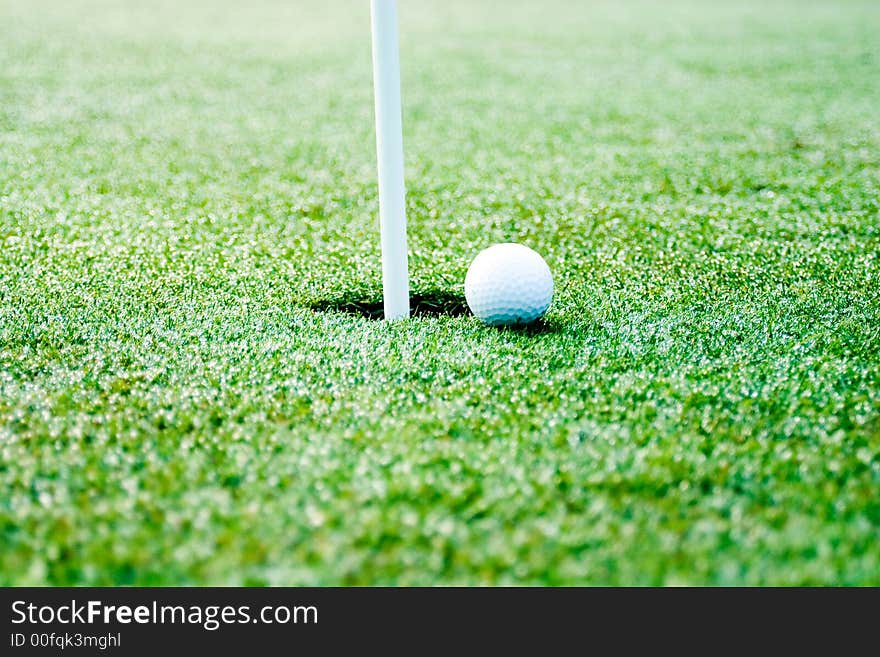 This is a picture of a golf ball next to the hole with flag pole showing. slective focus is on the ball and hole. This is a picture of a golf ball next to the hole with flag pole showing. slective focus is on the ball and hole.