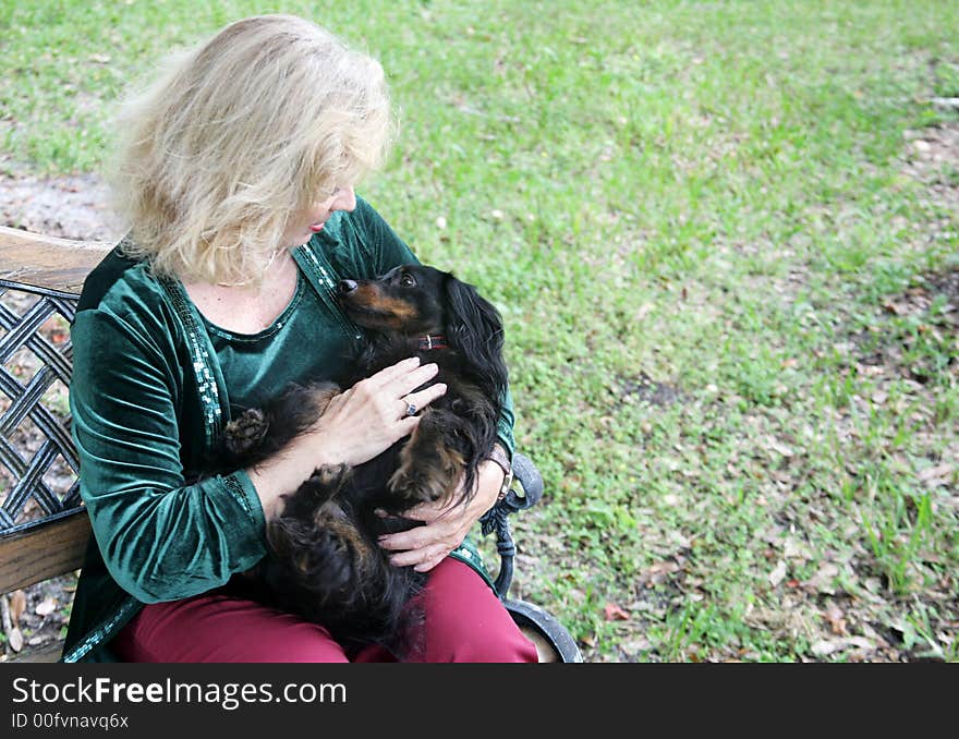 A blond woman sitting on a park bench holding her dachshund like a baby and petting him. Room for text. A blond woman sitting on a park bench holding her dachshund like a baby and petting him. Room for text.