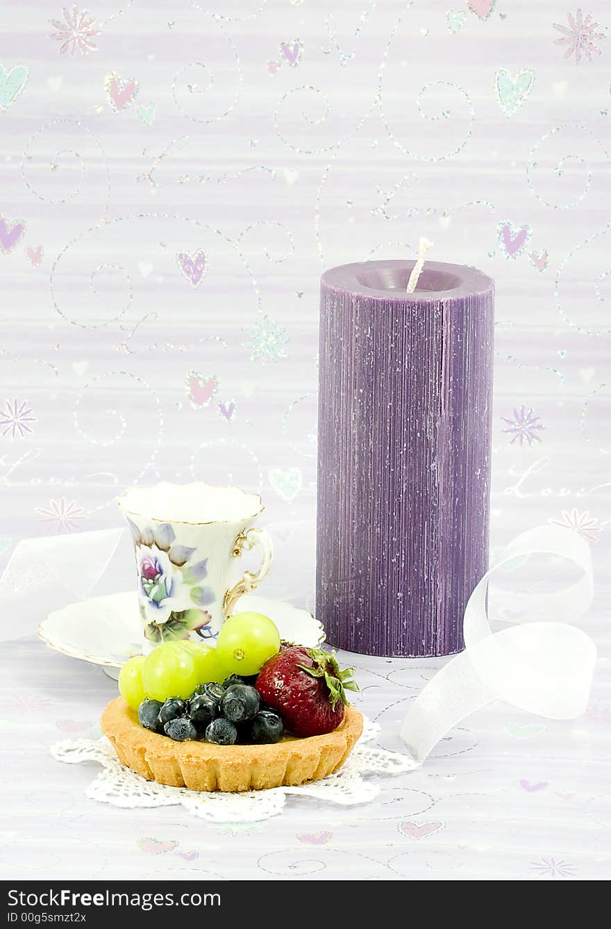 Fruit tart covered with strawberries,blueberries and green grapes on a doily with purple candle and a vintage tea cup and delicate white ribbon. Fruit tart covered with strawberries,blueberries and green grapes on a doily with purple candle and a vintage tea cup and delicate white ribbon.