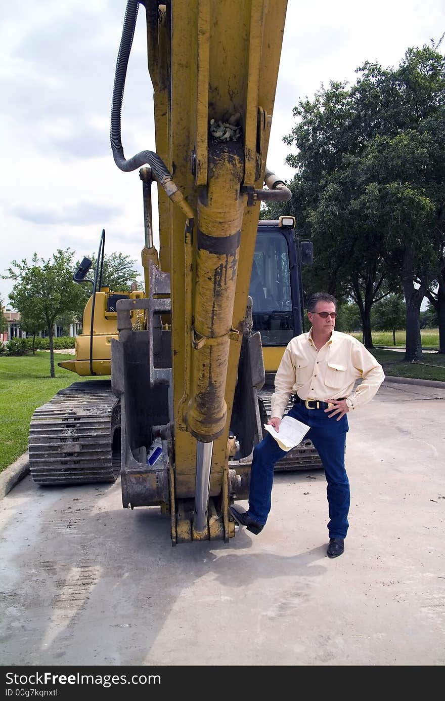 A man standing with his foot on the bucket of an idle backhoe with documents in his hand. A man standing with his foot on the bucket of an idle backhoe with documents in his hand.