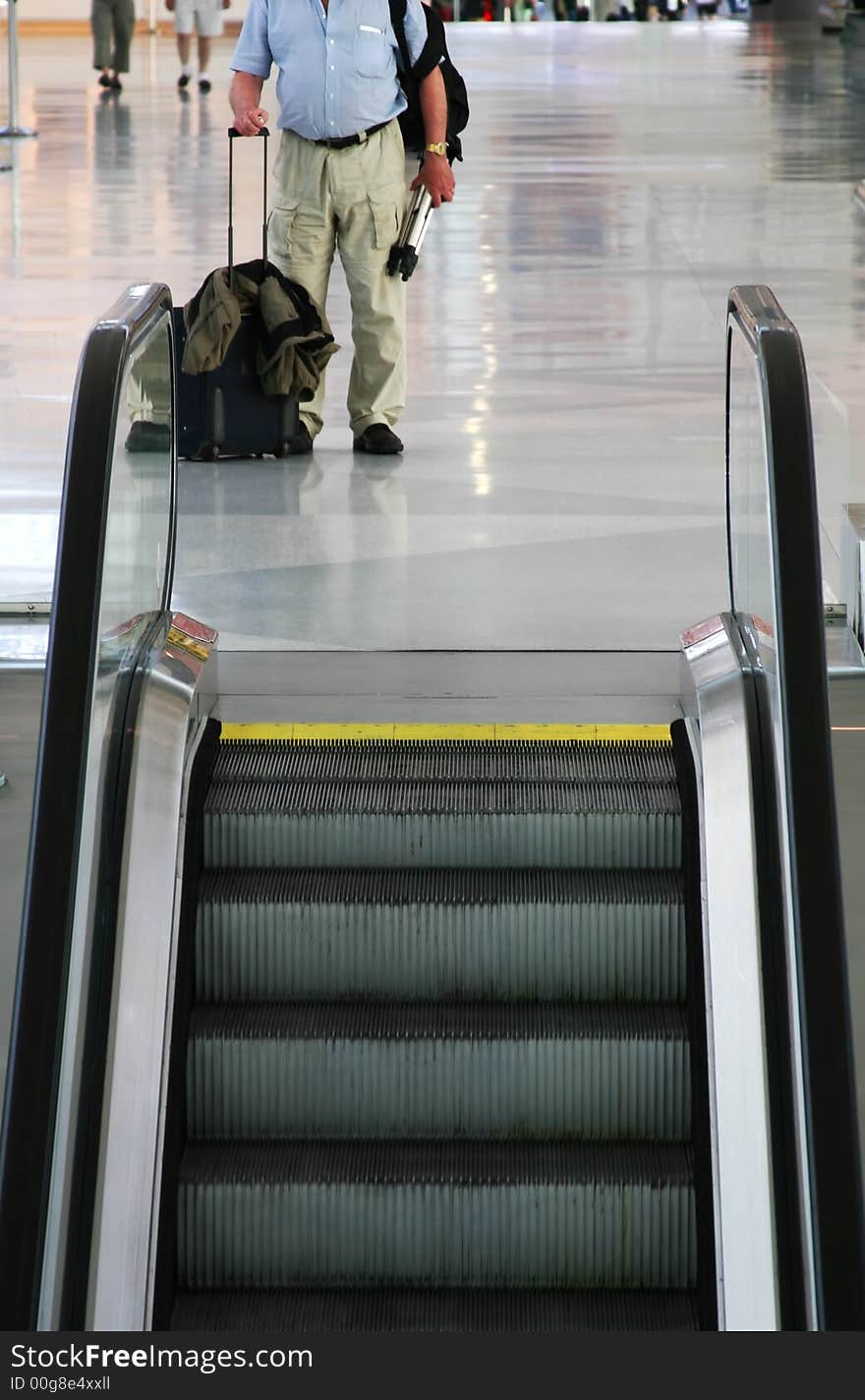 A bodyshot of a man standing by escalators in an airport with hand luggage. A bodyshot of a man standing by escalators in an airport with hand luggage