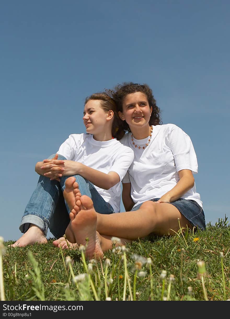 Two nice young girls sit on a grass on a background of the blue sky, smile and wave hands. Two nice young girls sit on a grass on a background of the blue sky, smile and wave hands