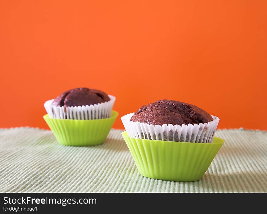 Two muffins in lime green plastic cups against orange wall on cotton table cloth. Focus on first muffin. Two muffins in lime green plastic cups against orange wall on cotton table cloth. Focus on first muffin