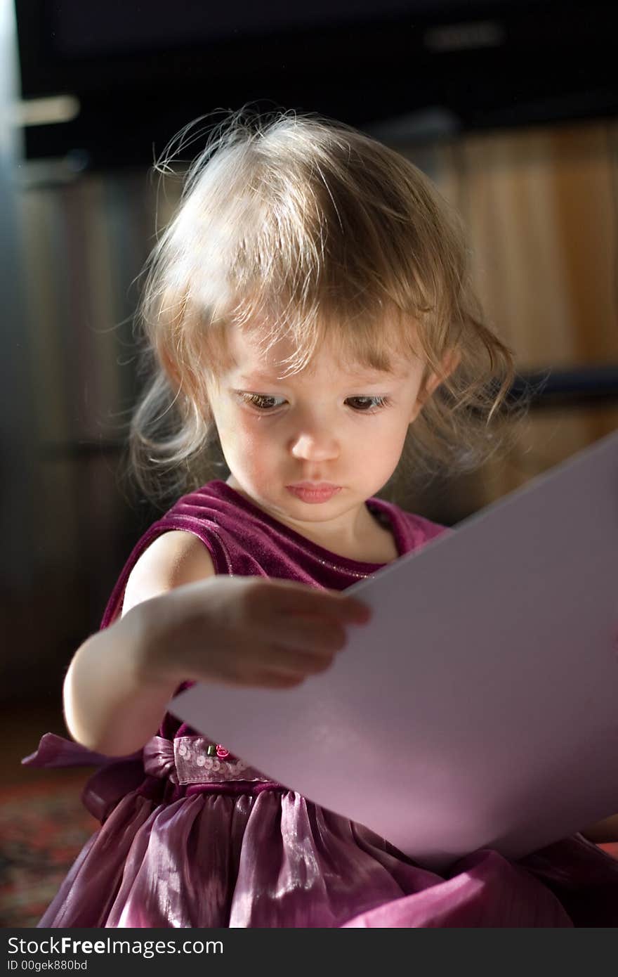 Beautiful small girl is sitting in the rays of sun and looking into the book