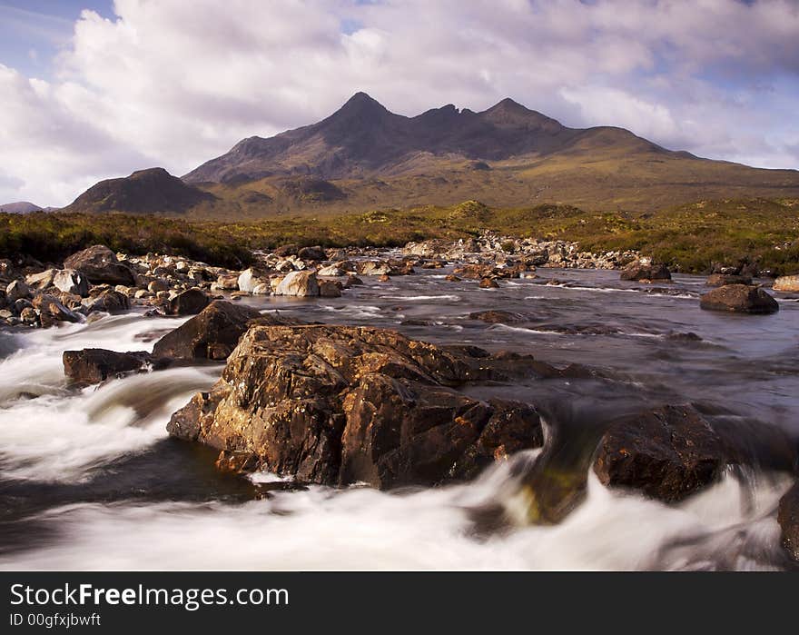 Morning sunlight lights the distant cullin hills in a warm glow. The photo is taken over a raging mountain stream with large rocks dominating the foreground. Morning sunlight lights the distant cullin hills in a warm glow. The photo is taken over a raging mountain stream with large rocks dominating the foreground