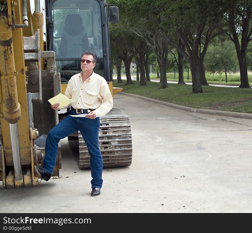 A man standing with his foot on the bucket of an idle backhoe with documents in his hand. A man standing with his foot on the bucket of an idle backhoe with documents in his hand.