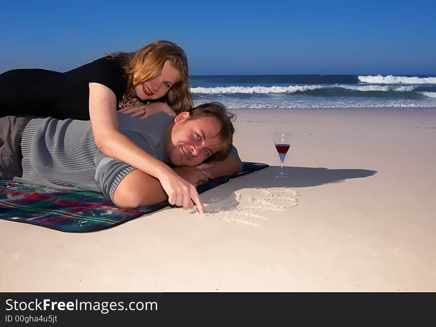 Young married couple enjoying their time on the beach. The lady is lying on her husband's back and drawing on the sand. Young married couple enjoying their time on the beach. The lady is lying on her husband's back and drawing on the sand.