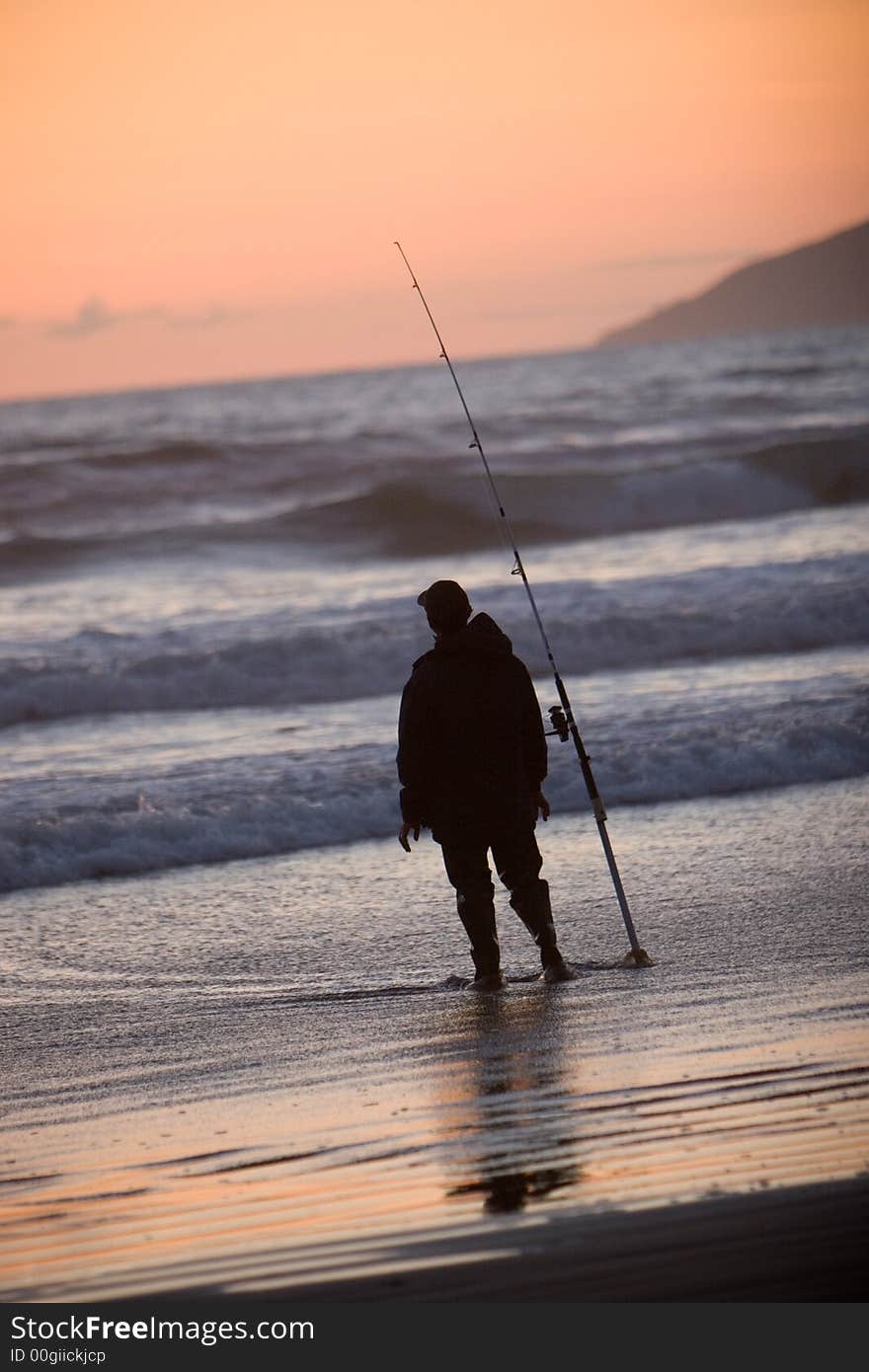 Silhouette of man fishing in the ocean from the beach at sunset.