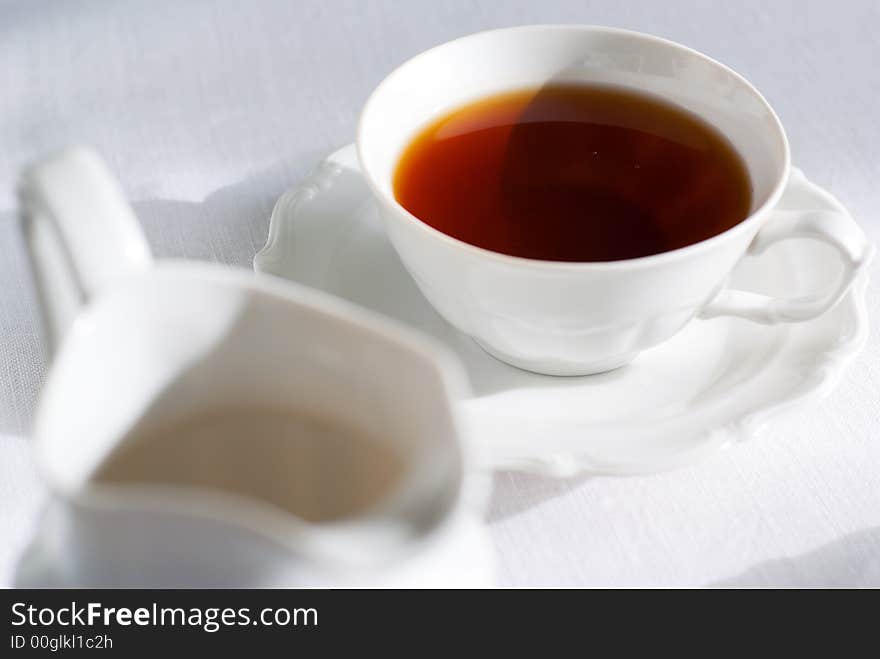 Cup of tea (focus on it) and porcelain milk jug on the white tablecloth. Shallow depth of field. Cup of tea (focus on it) and porcelain milk jug on the white tablecloth. Shallow depth of field.