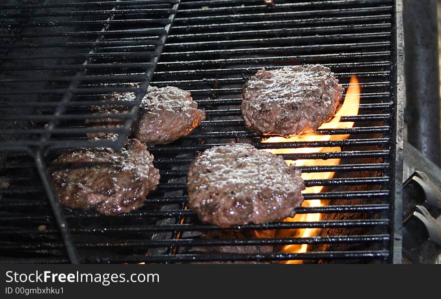 Hamburgers sit cooking with fire on a grill . Hamburgers sit cooking with fire on a grill .