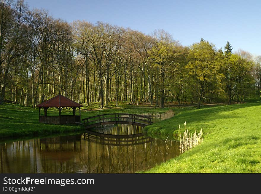 small wooden bridge and alcove in the park with fresh green trees, grass on the blue sky. small wooden bridge and alcove in the park with fresh green trees, grass on the blue sky