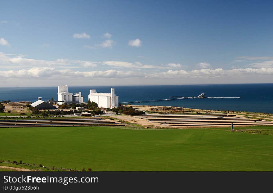 Loading site with white tower silo and jetty. Sea and blue sky in the background, and green field in foreground. Loading site with white tower silo and jetty. Sea and blue sky in the background, and green field in foreground.