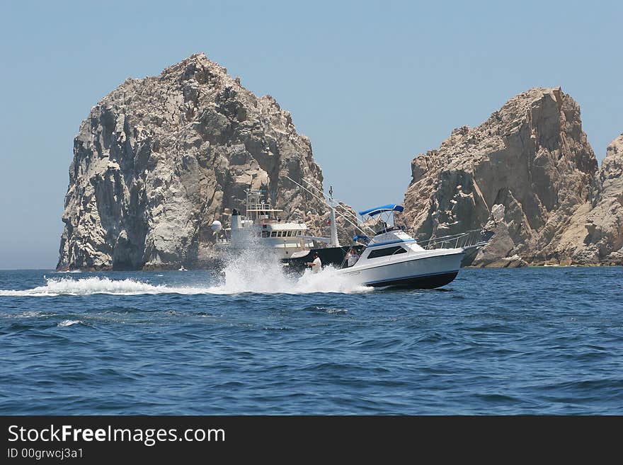 Fast cabin cruiser speeding past anchored boat at rocks. Fast cabin cruiser speeding past anchored boat at rocks