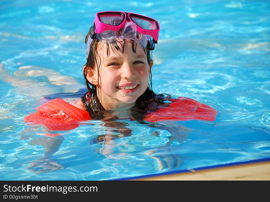 A young girl smiles into the camera as she takes a dip in the pool on a sunny day. She is wearing floats and goggles. A young girl smiles into the camera as she takes a dip in the pool on a sunny day. She is wearing floats and goggles.