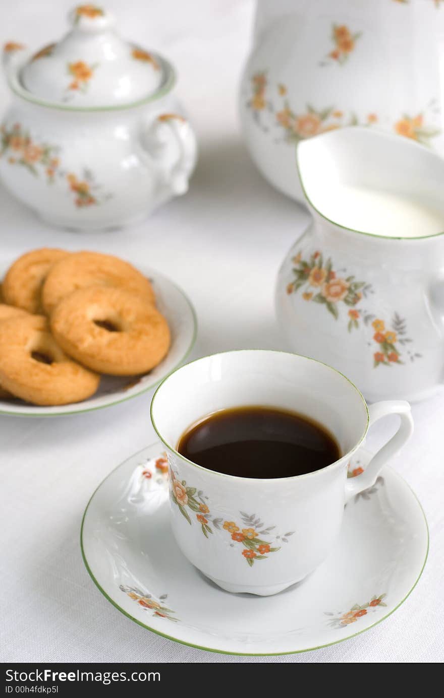 Coffee (or tea) set on the table - cup of coffee on the front (focus on it) cookies, milk jug and sugar bowl behind. Coffee (or tea) set on the table - cup of coffee on the front (focus on it) cookies, milk jug and sugar bowl behind.