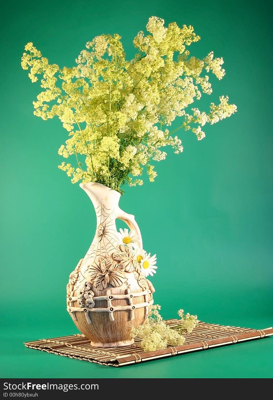 On a wooden napkin a jug with bouquets of yellow field colors cost. It is photographed on a green background. On a wooden napkin a jug with bouquets of yellow field colors cost. It is photographed on a green background.