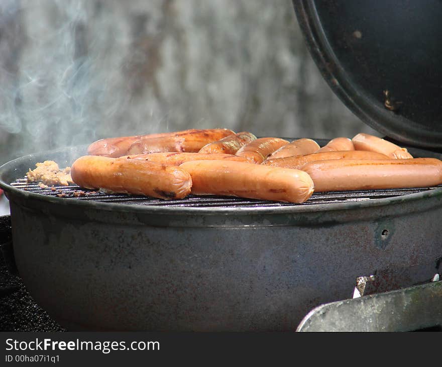 Lots of hot dogs cooking on a old grill with smoke rising.