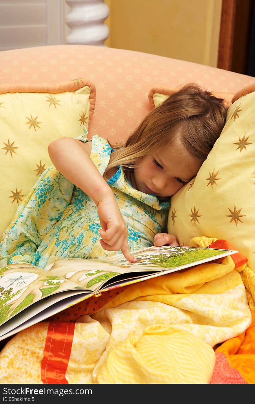 Adorable little girl looking at a book in a chair with a quuilt over her. Adorable little girl looking at a book in a chair with a quuilt over her