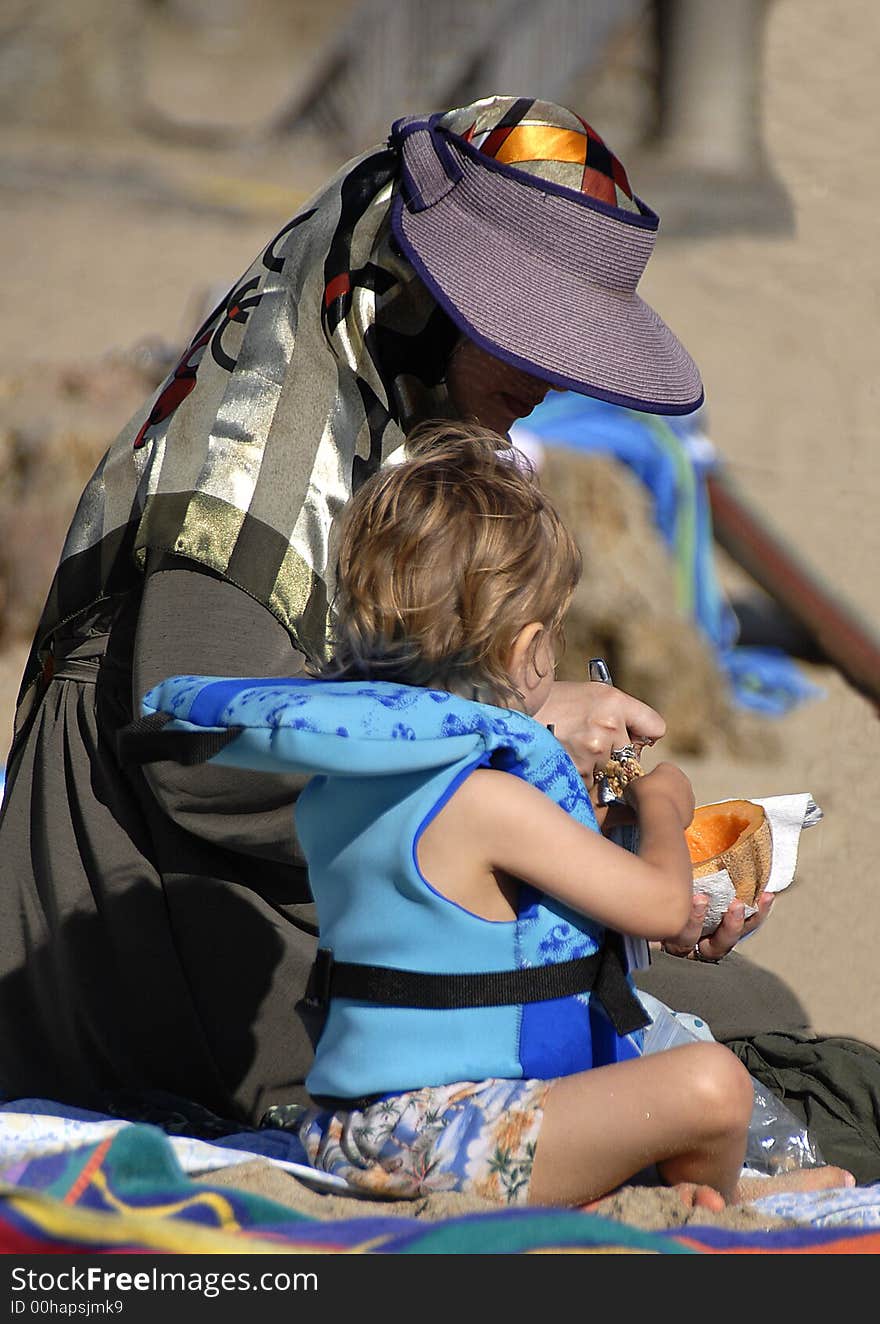 A mom and a child are eating on the beach. A mom and a child are eating on the beach