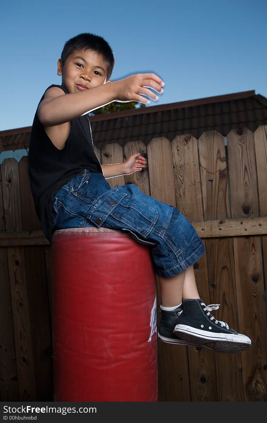 Young asian boy sitting on top of a punching bag outside beside a tall wooden fence smiling wearing jeans and black tshirt. Young asian boy sitting on top of a punching bag outside beside a tall wooden fence smiling wearing jeans and black tshirt