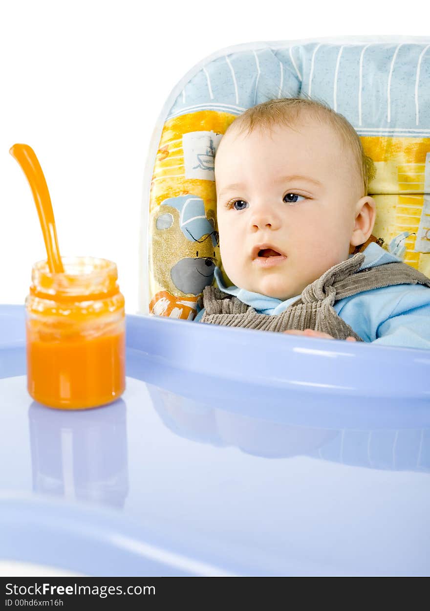 Pretty infant sitting on baby's chair. Looking at orange pulp in jar. White background. Pretty infant sitting on baby's chair. Looking at orange pulp in jar. White background