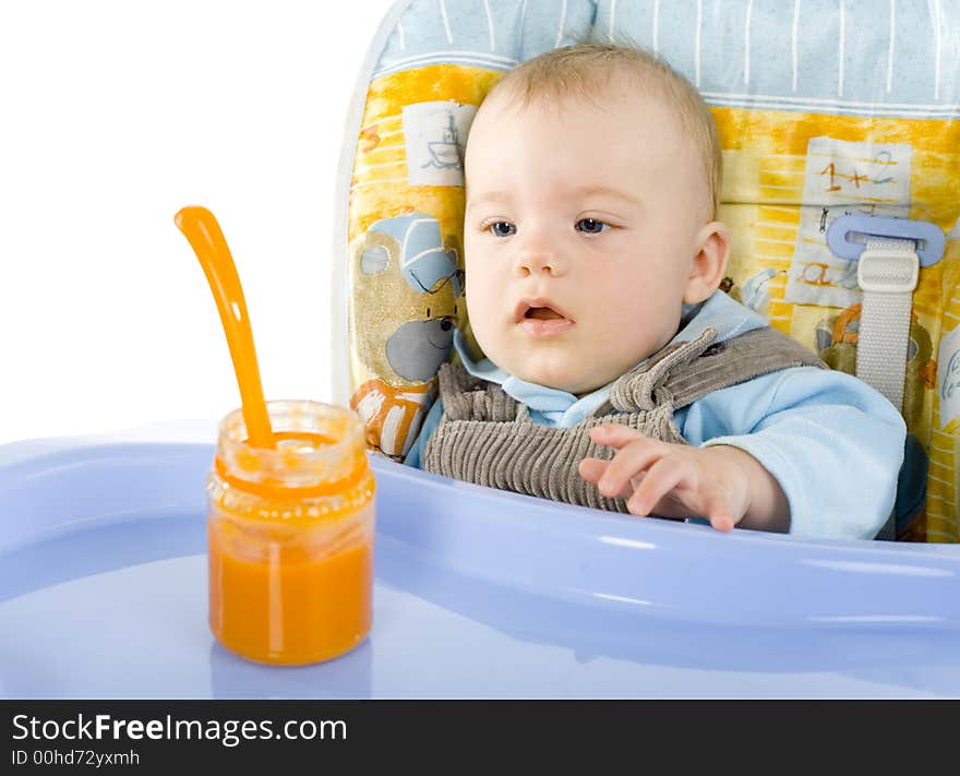 Pretty infant sitting on baby's chair. Looking at orange pulp in jar. White background. Pretty infant sitting on baby's chair. Looking at orange pulp in jar. White background