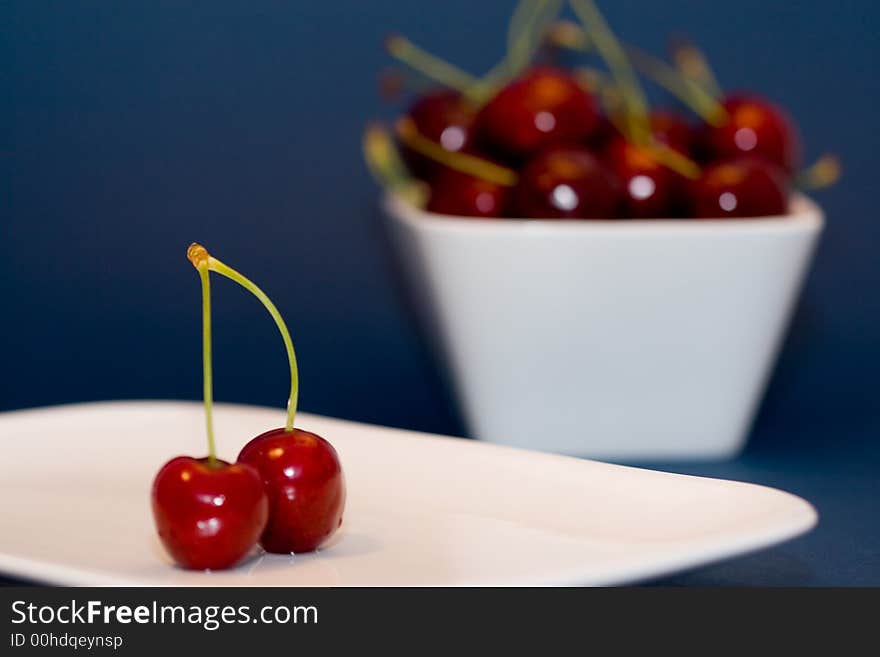 Multiple moist red cherries in a white bowl and 2 cherries in front. Multiple moist red cherries in a white bowl and 2 cherries in front