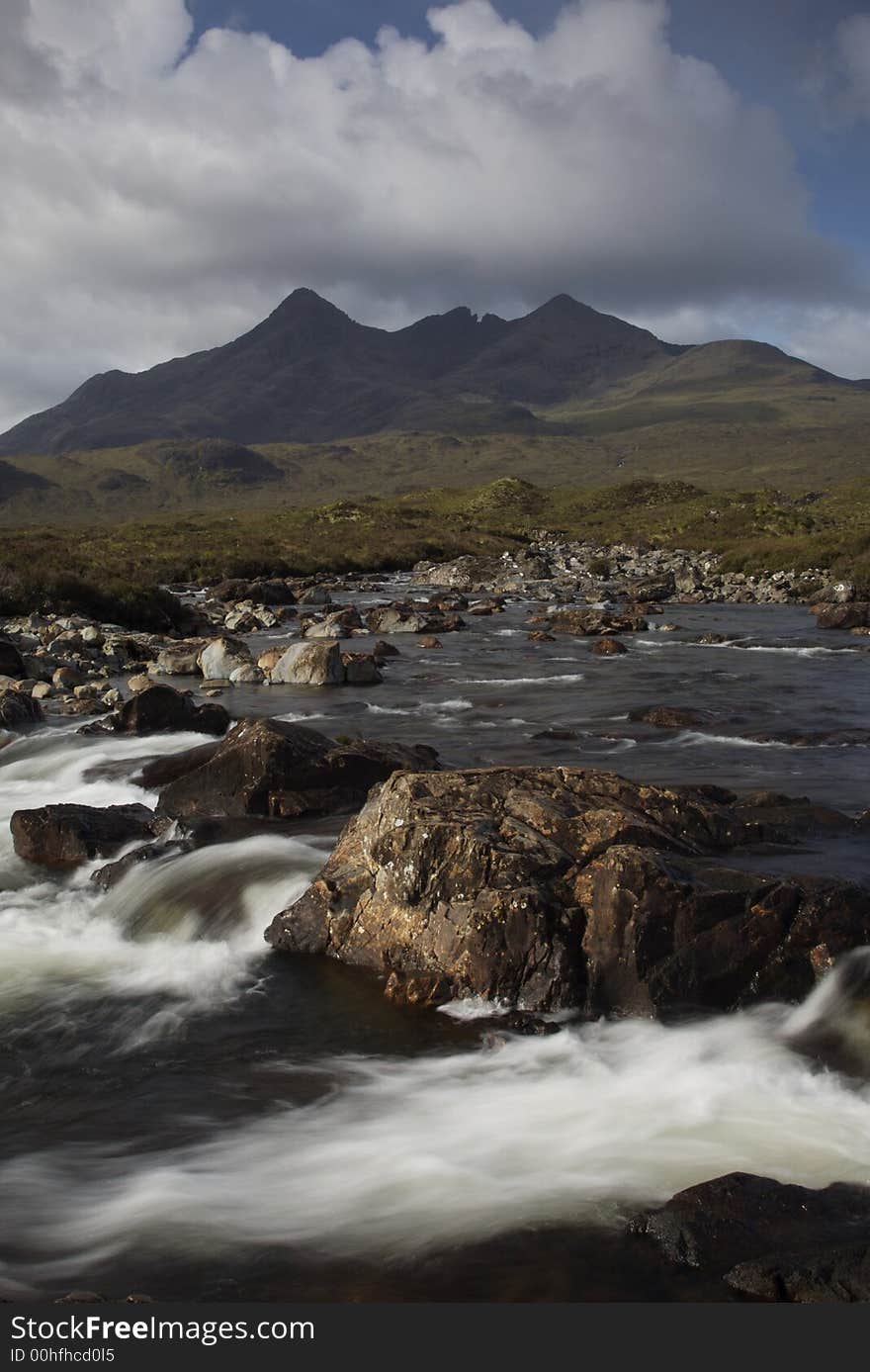 Morning sunlight lights the distant cullin hills in a warm glow. The photo is taken over a raging mountain stream with large rocks dominating the foreground. Morning sunlight lights the distant cullin hills in a warm glow. The photo is taken over a raging mountain stream with large rocks dominating the foreground