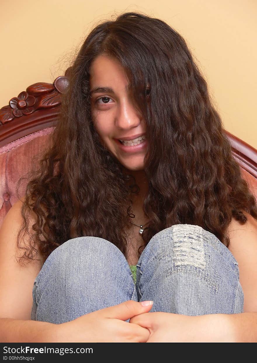 A happy girl, sitting on the sofa her hands around her knees and her long dark curly hair is coffering half her face.