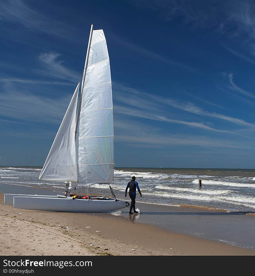 A catamaran sail boat at the beach with white sail and beautiful blue sky and some white clouds. A catamaran sail boat at the beach with white sail and beautiful blue sky and some white clouds.