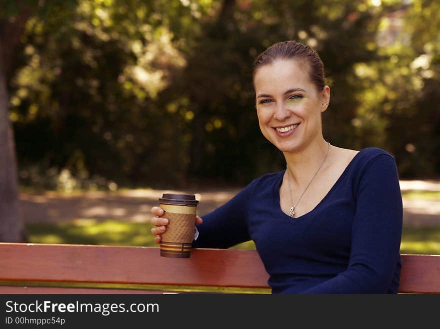 A smiling, beautiful young woman relaxing on park bench with a cup of coffee or tea. A smiling, beautiful young woman relaxing on park bench with a cup of coffee or tea.