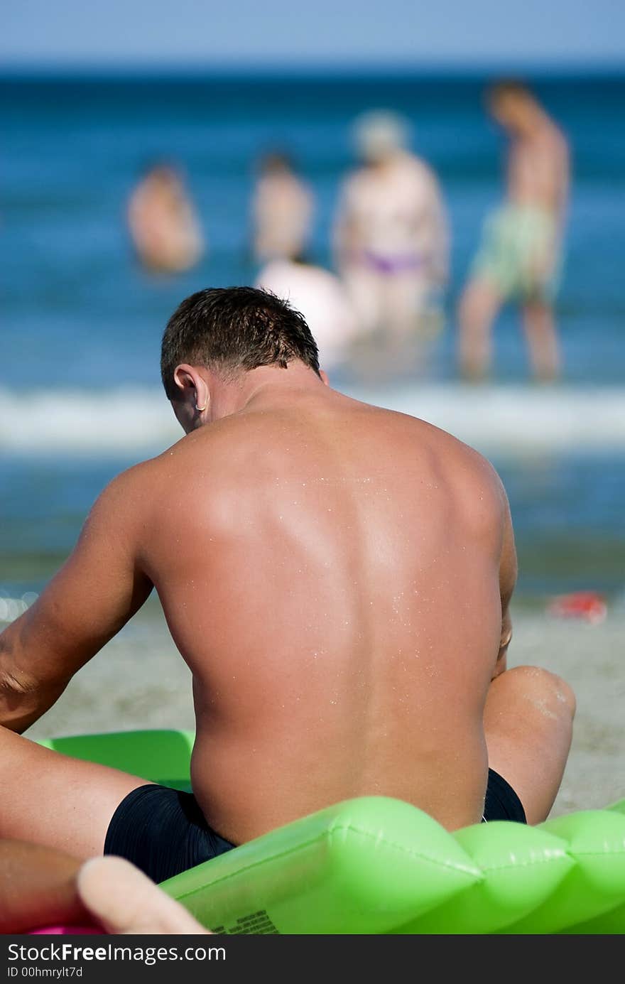 Man resting and sunbathing on the beach, people and sea in the background.