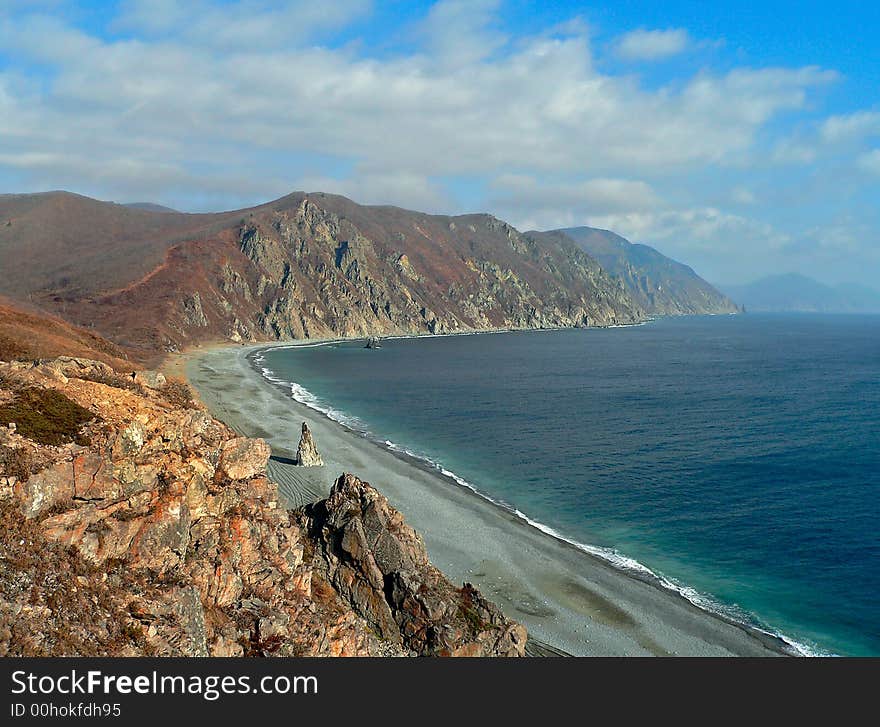 A cloudscape on sea: turquoise sea, grey beach, yellow-red capes and rocks and white clouds on azure sky. Autumn. Russian Far East, Primorye, Japanese sea, Tasovaya bay. A cloudscape on sea: turquoise sea, grey beach, yellow-red capes and rocks and white clouds on azure sky. Autumn. Russian Far East, Primorye, Japanese sea, Tasovaya bay.