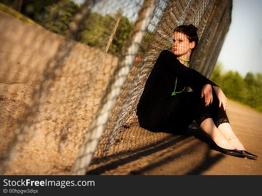 Attractive young lady sitting against a fence with a chain-link shadow criss-crossing her face. Attractive young lady sitting against a fence with a chain-link shadow criss-crossing her face.