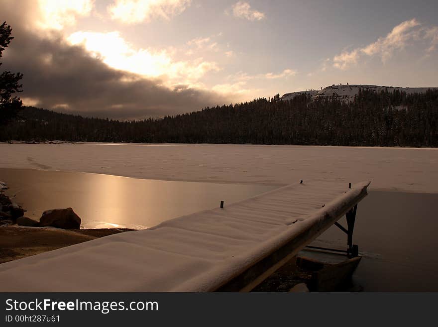 A snow covered dock leads to an iceed over lake as the sunrises over a near by mountain. A snow covered dock leads to an iceed over lake as the sunrises over a near by mountain