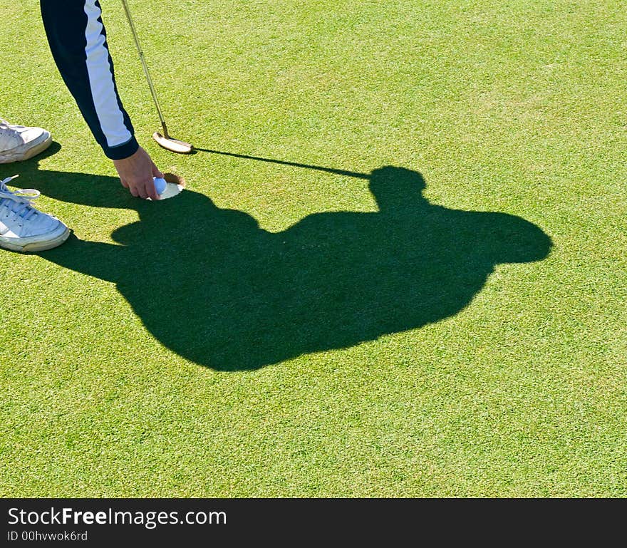 Shadow of a golfer taking the ball out of the cup after sinking a putt. Shadow of a golfer taking the ball out of the cup after sinking a putt