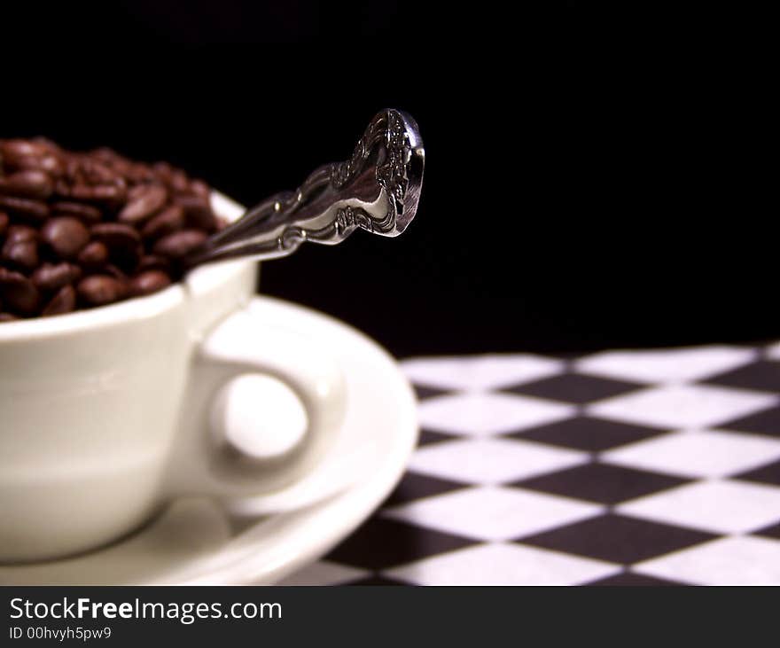 Color photo of a cup full of dark brown coffee beans with a silver spoon sticking out from it. The focus is on the tip of the spoon. Color photo of a cup full of dark brown coffee beans with a silver spoon sticking out from it. The focus is on the tip of the spoon.