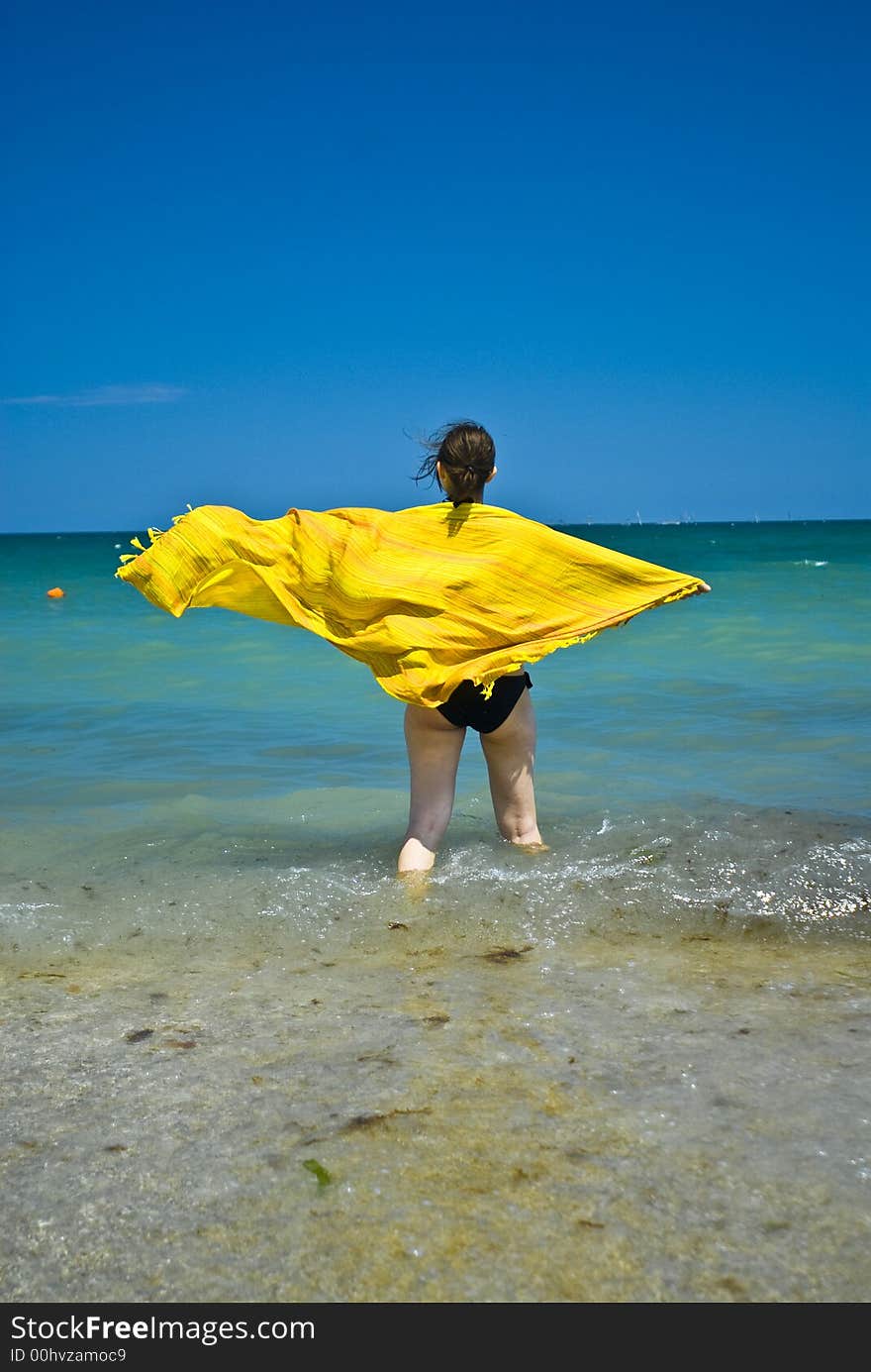 Young woman wrapped in colourful scarf blown by the summer breeze, wading in the shoreline shallow water.