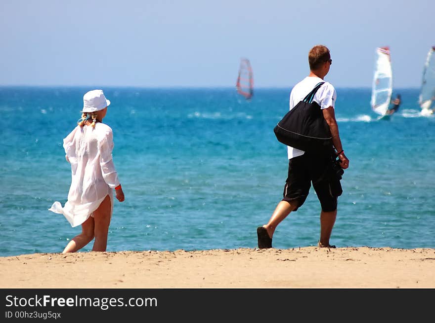 Young couple walking along a sandy beach, sunny day, summer season, in background wind surfers surfing in a blue sea. Young couple walking along a sandy beach, sunny day, summer season, in background wind surfers surfing in a blue sea.