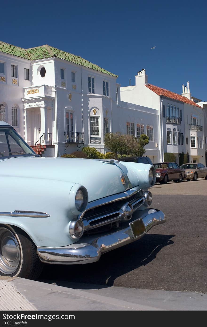 Front of a blue vintage car, with old white houses in the back