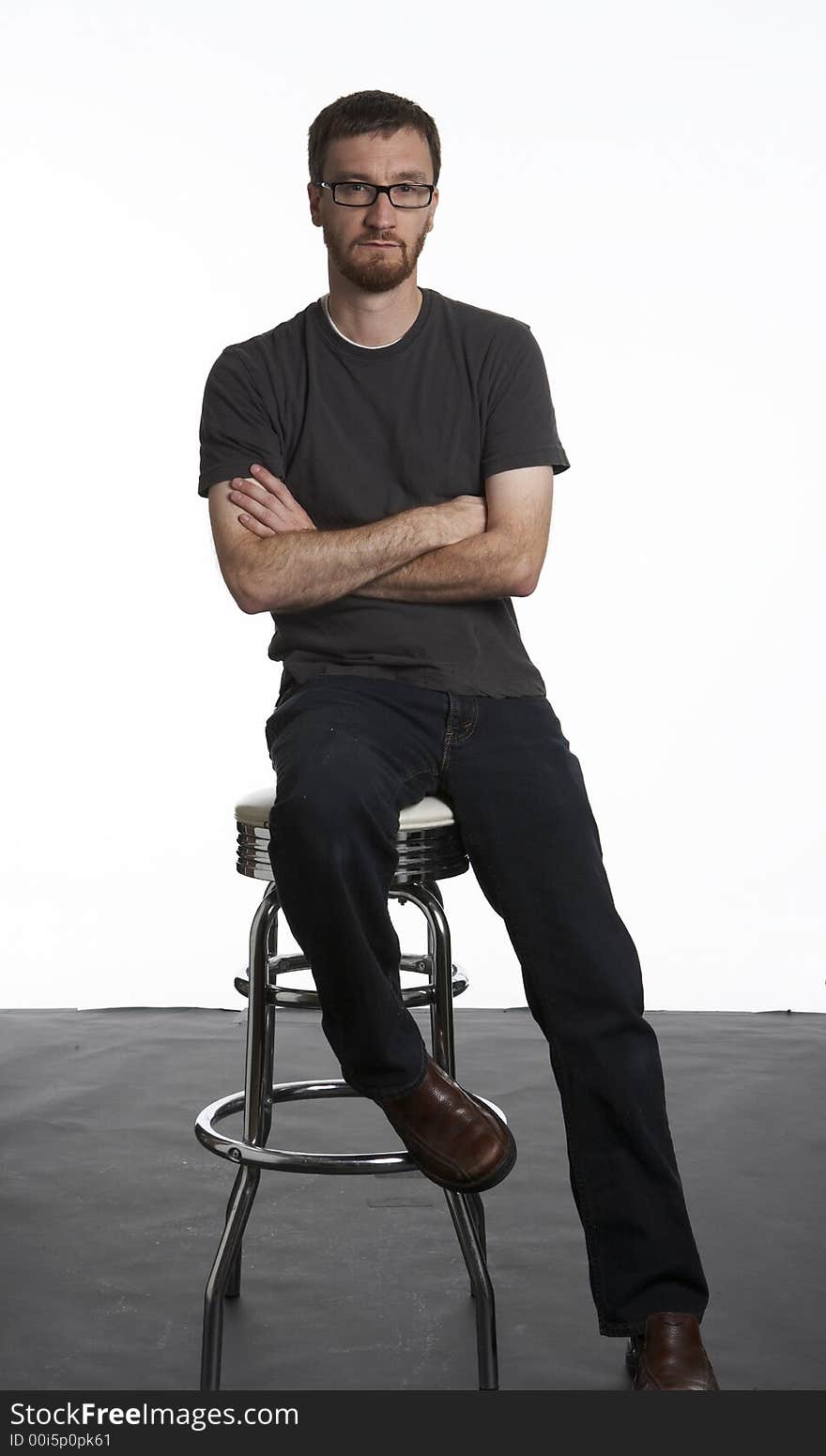 Portrait of a bearded man inside a studio. His arms are folded and he is sitting on a chrome stool. Portrait of a bearded man inside a studio. His arms are folded and he is sitting on a chrome stool