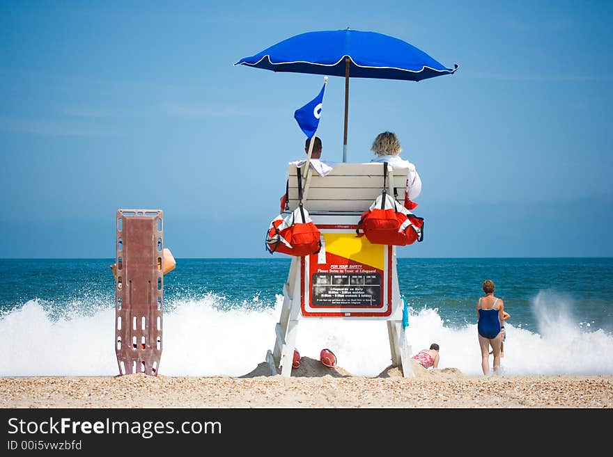 Two young men beach lifeguards watch people swimming in the blue ocean with waves crashing on a sandy beach. Room for text. Two young men beach lifeguards watch people swimming in the blue ocean with waves crashing on a sandy beach. Room for text.