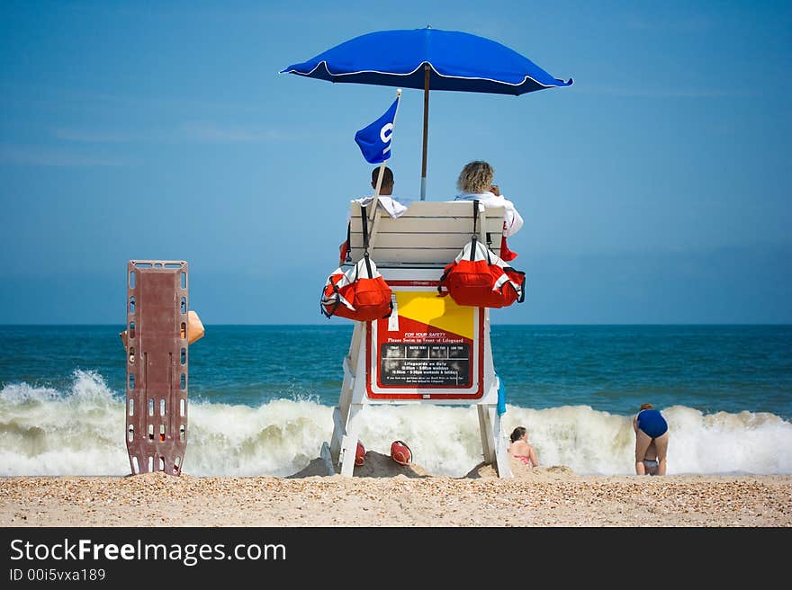 Two young men beach lifeguards watch people swimming in the blue ocean with waves crashing on a sandy beach. Room for text. Two young men beach lifeguards watch people swimming in the blue ocean with waves crashing on a sandy beach. Room for text.