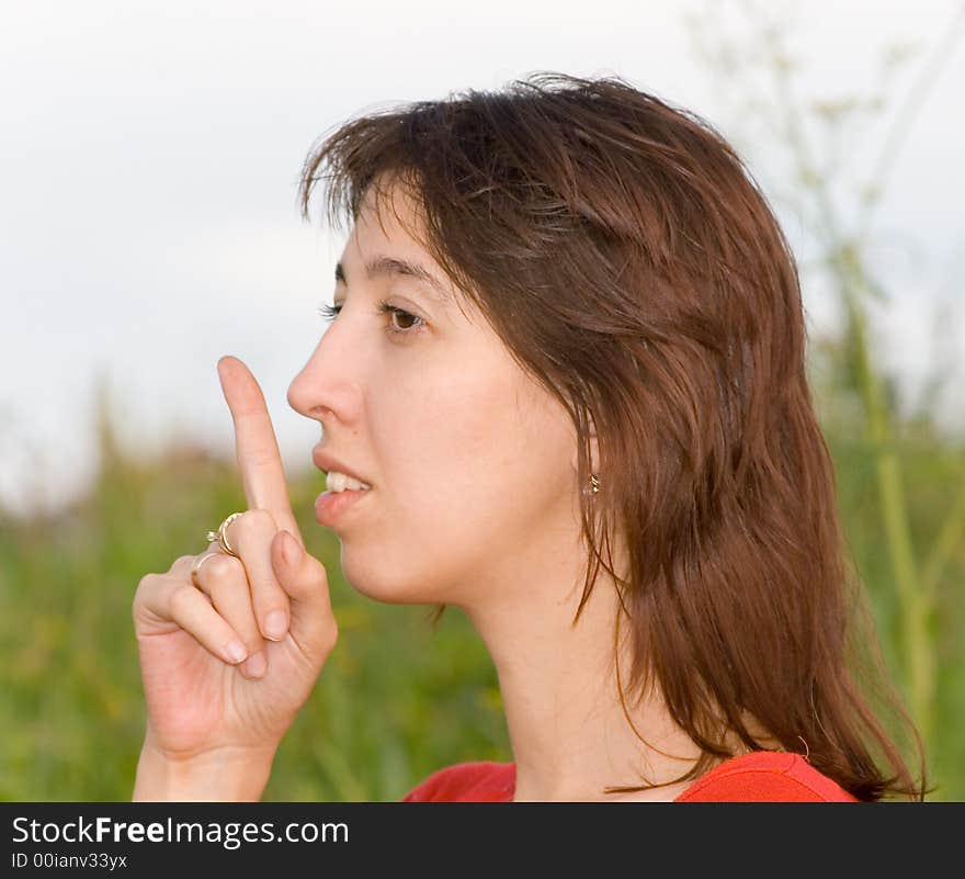 Photo of the young woman in a structure, calling for silence gesture (a finger enclosed to lips) on a background of the blue sky with clouds and a green grass. Focus of a photo on eyes of the woman