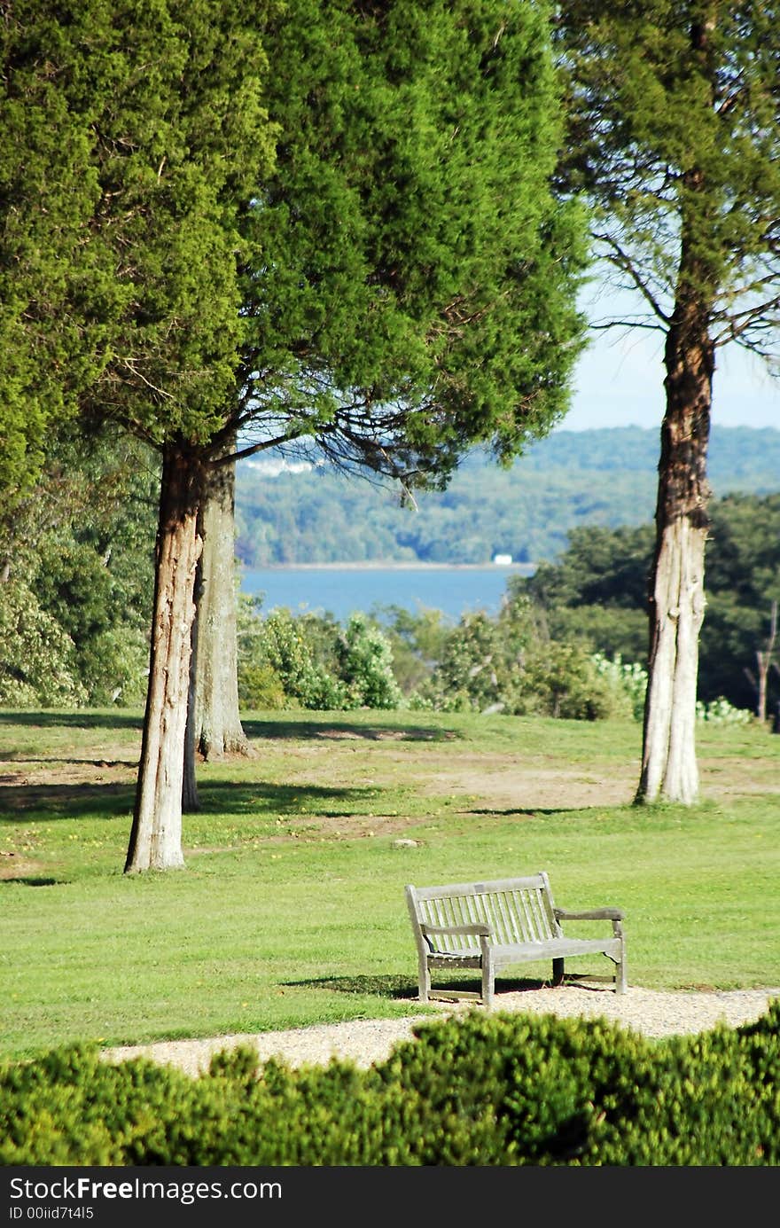 A vacant bench shownn on clear warm day in spring, summer, or early fall with trees, grass, greenry with blue water in background. A vacant bench shownn on clear warm day in spring, summer, or early fall with trees, grass, greenry with blue water in background