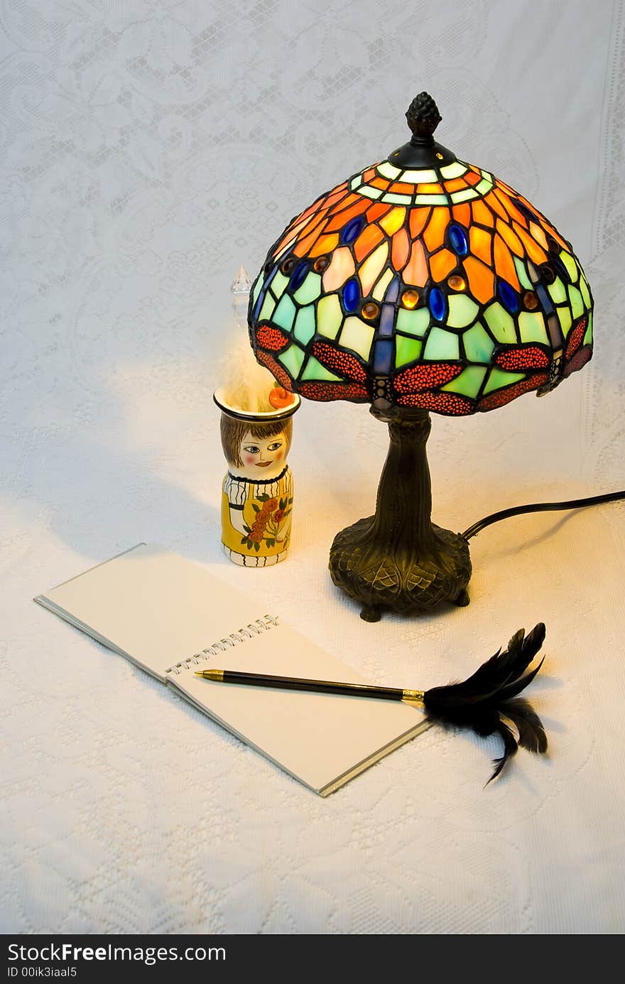 Vertical view of lit stained glass style lamp with open notepad and black pen with feathers. Vertical view of lit stained glass style lamp with open notepad and black pen with feathers