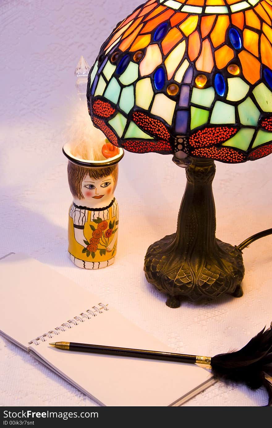 Vertical view of lit stained glass style lamp with notepad, black pen with feather and ceramic pencil holder. Vertical view of lit stained glass style lamp with notepad, black pen with feather and ceramic pencil holder