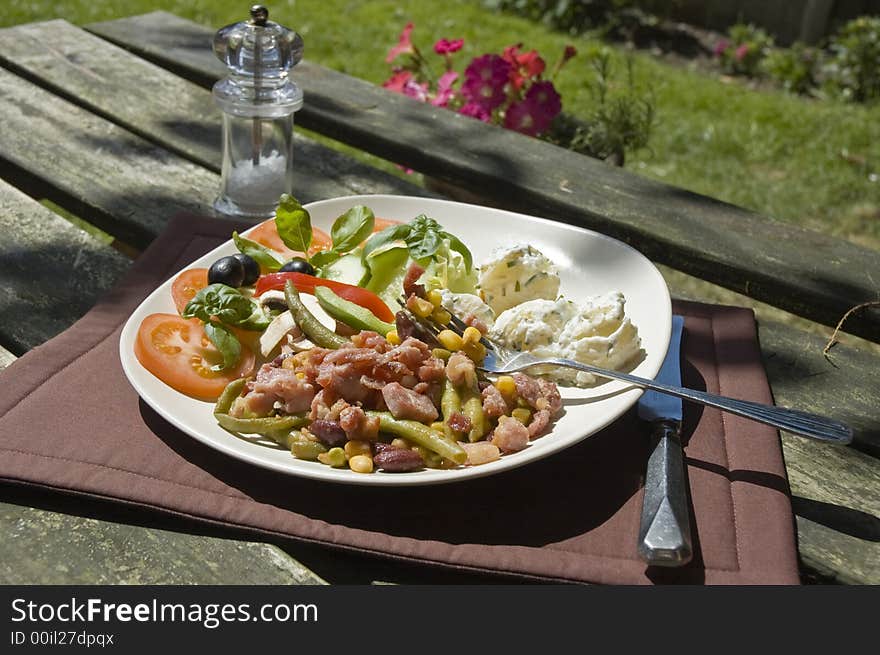 A garden scene with a plate of mixed prepared salads.  Potatoe & chive, Bean salad topped with crispy bacon, and italian style salad with peppers, tomatoes fresh basil & calamata olives. A garden scene with a plate of mixed prepared salads.  Potatoe & chive, Bean salad topped with crispy bacon, and italian style salad with peppers, tomatoes fresh basil & calamata olives