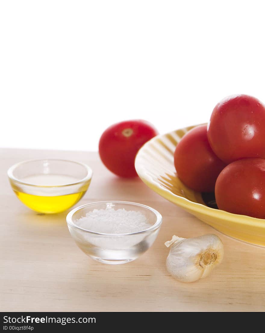 A portion of olive oil, small portion of salt, garlic clove, and a yellow plate with tomatoes on top of a wooden cutting board against a white background. A portion of olive oil, small portion of salt, garlic clove, and a yellow plate with tomatoes on top of a wooden cutting board against a white background.